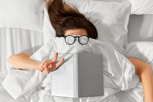 people, bedtime and rest concept - woman lying in bed under white blanket or duvet with book and glasses showing peace hand sign
