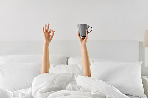 morning, comfort and people concept - young woman with cup of coffee lying in bed and showing ok hand sign at home bedroom