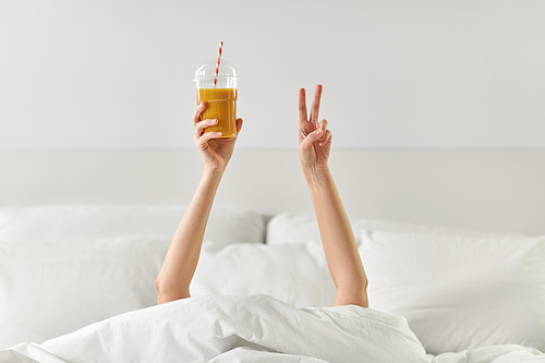 drink, comfort and morning concept - hands of young woman lying in bed with cup of juice and showing peace hand sign at bedroom