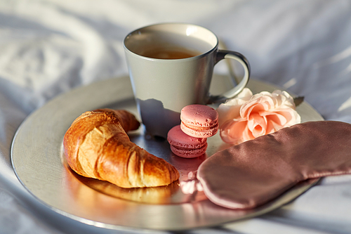 morning, hygge and breakfast concept - croissant, cup of coffee, macaroons and eye sleeping mask on plate in bed at home