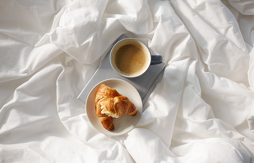 morning, hygge and breakfast concept - croissants, cup of coffee and book in bed at home