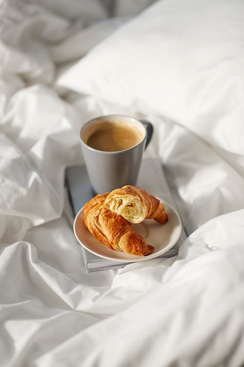morning, hygge and breakfast concept - croissants, cup of coffee and book in bed at home