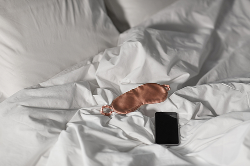 hygge and nighttime concept - smartphone and eye sleeping mask in bed at night