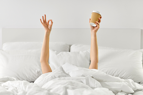 morning, comfort and people concept - young woman with cup of takeaway coffee lying in bed and showing ok hand sign at home bedroom