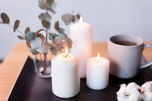 decoration, hygge and cosiness concept - burning white candles, tea in mug, branches of eucalyptus populus and cotton flowers on table