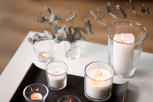 decoration, hygge and cosiness concept - burning white fragrance candles on tray and branches of eucalyptus populus on table at cozy home