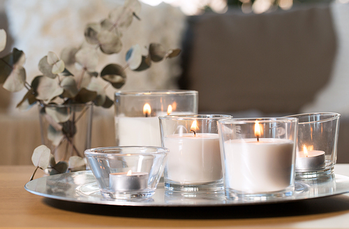 decoration, hygge and cosiness concept - burning white fragrance candles on tray and branches of eucalyptus populus on table at cozy home