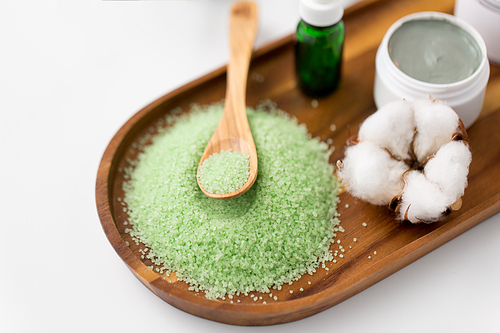 beauty, spa and wellness concept - green bath salt with wooden spoon, blue clay mask, cotton flower and serum on tray