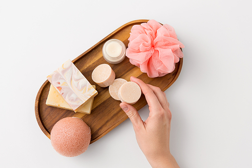 beauty, spa and wellness concept - close up of hand with crafted soap bars, konjac sponge, moisturizer and wisp on wooden tray