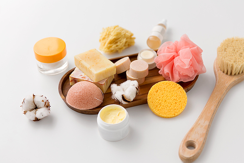 beauty and spa concept - close up of konjac sponge, crafted soap bars, body butter, wisp and natural bristle bath brush and moisturizer on wooden tray