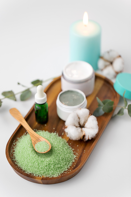 beauty and spa concept - green bath salt, serum with dropper, blue clay mask, moisturizer and eucalyptus cinerea with cotton flowers on wooden tray