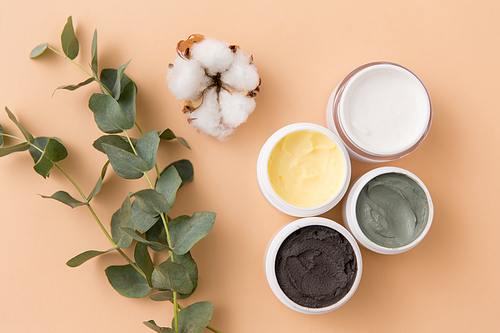beauty, spa and wellness concept - blue clay mask, body butter, therapeutic mud and eucalyptus cinerea with cotton flower on beige background