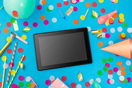 celebration, holidays and technology concept - black tablet computer, birthday party props and colorful confetti on blue background