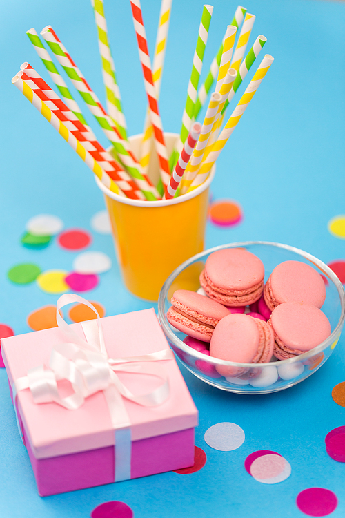 birthday party, celebration and decoration concept - gift box, pink macarons, paper straws and confetti on blue background