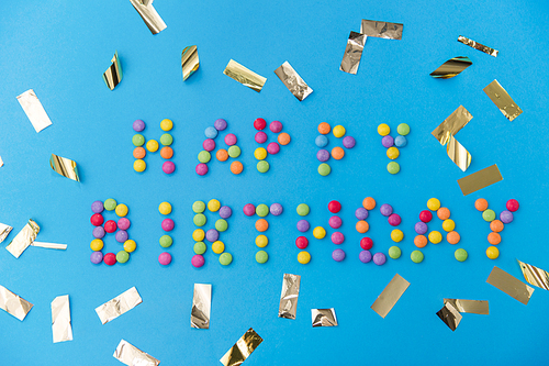 food, confectionery and sweets concept - happy birthday lettering made of candy drops and metallic confetti on blue background