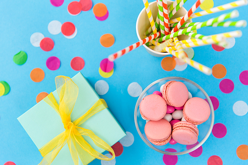birthday party, celebration and decoration concept - gift box, pink macarons, paper straws and confetti on blue background