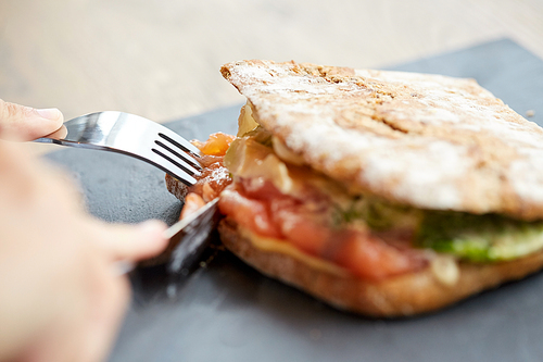 food, dinner and people concept - hand of person eating salmon panini sandwich with tomatoes and cheese using fork and knife at restaurant