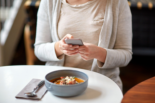 food, new nordic cuisine, technology, eating and people concept - woman sitting at cafe table with smartphone and bowl of vegetable pumpkin-ginger soup with goat cheese and tomato salad with yogurt
