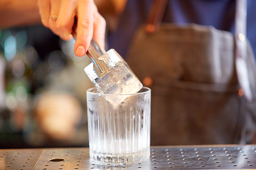 alcohol drinks, people and luxury concept - bartender with tongs adding ice cube into glass and preparing cocktail at bar counter