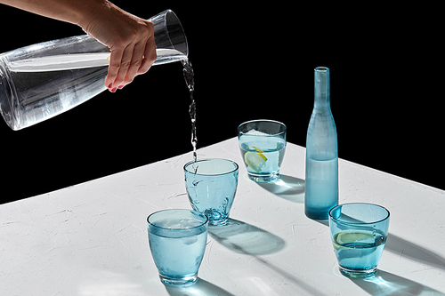 drink, detox and diet concept - hand pouring water to blue glasses with lemons and ice on white table over black background