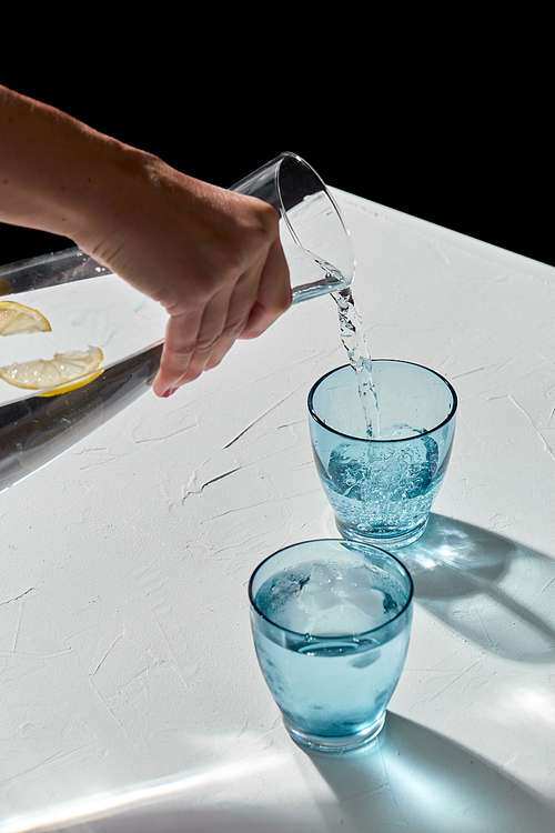 drink, detox and diet concept - hand pouring water to blue glasses with lemons and ice on white table over black background