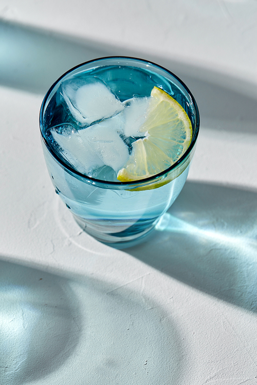 drink, detox and diet concept - blue glass of water with lemon and ice on white background
