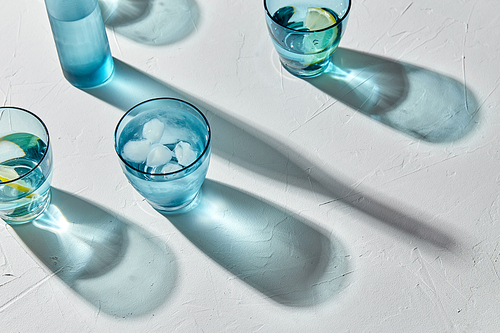 drink, detox and diet concept - blue glasses with water, lemons and ice on white background