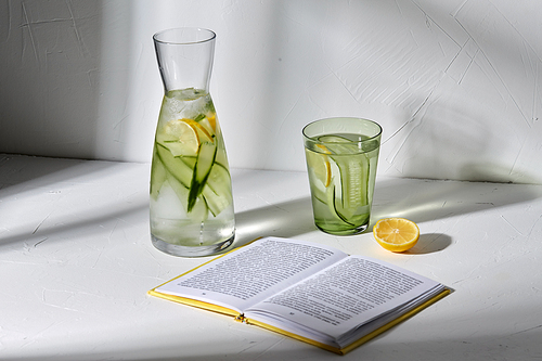 drink, detox and diet concept - glasses with fruit water with lemon and cucumber and open book dropping shadows on white surface