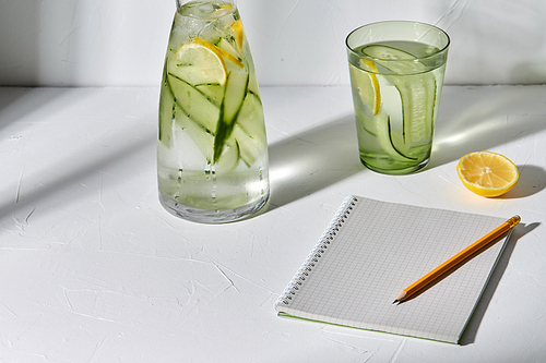 drink, detox and diet concept - glasses with fruit water with lemon and cucumber and notebook with pencil dropping shadows on white surface
