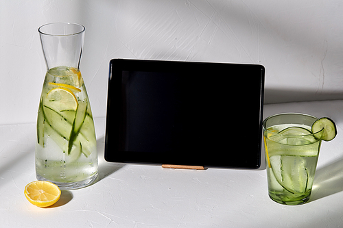 drink, detox and diet concept - glasses with fruit water with lemon and cucumber and notebook with tablet pc computer on white table