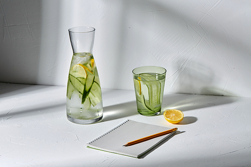 drink, detox and diet concept - glasses with fruit water with lemon and cucumber and notebook with pencil dropping shadows on white surface