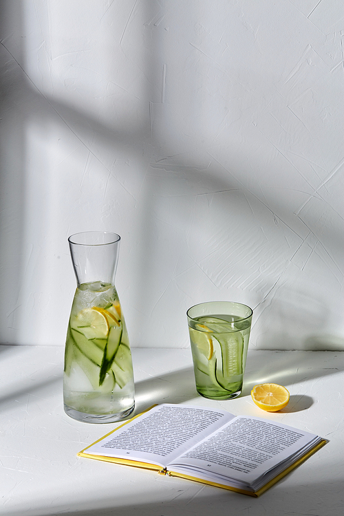 drink, detox and diet concept - glasses with fruit water with lemon and cucumber and open book dropping shadows on white surface
