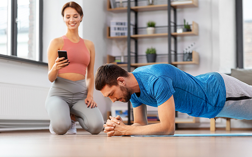 sport, fitness, lifestyle and people concept - smiling young woman with smartphone and man exercising and doing plank at home