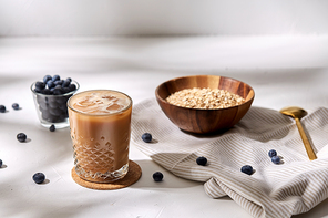 food, drinks and breakfast concept - glass of ice coffee on cork drink coaster, oatmeal in wooden bowl and spoon with blueberries on kitchen towel