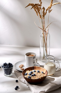 food, eating and breakfast concept - oatmeal in wooden bowl with blueberries, milk in glass jug and cup of coffee with brown sugar on kitchen towel