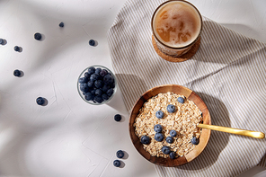 food, eating and breakfast concept - oatmeal in wooden bowl with blueberries and spoon, glass of ice coffee on cork drink coaster and kitchen towel