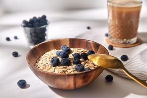food, healthy eating and breakfast concept - oatmeal in wooden bowl with blueberries and spoon, glass of ice coffee on cork drink coaster and kitchen towel