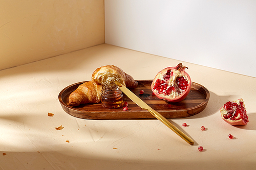 food and eating concept - croissant, pomegranate and honey with table knife on wooden tray