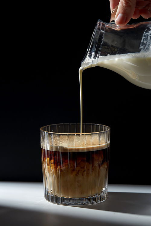 caffeine and drinks concept - hand with jug pouring cream or milk to glass of coffee on table
