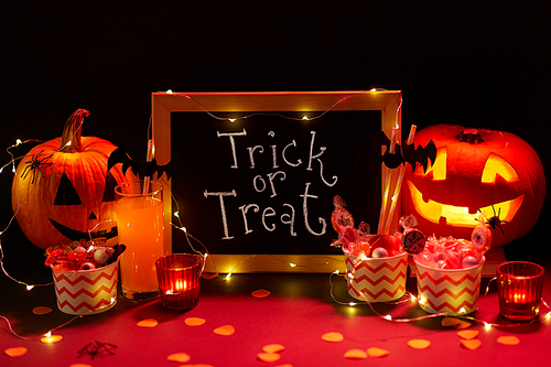 halloween and holiday decorations concept - chalkboard with trick or treat lettering, jack-o-lanterns or carved pumpkins, candies, burning candles and glass of juice with paper straw in dark room