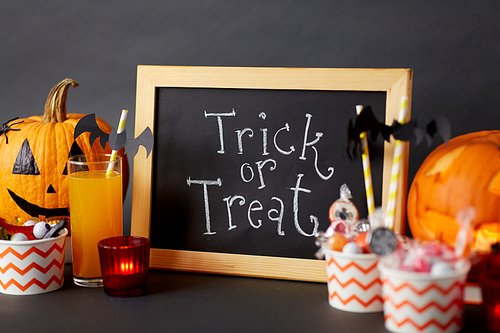 halloween and holiday decorations concept - chalkboard with trick or treat lettering, jack-o-lanterns or carved pumpkins, candies, burning candles and glass of juice with paper straw