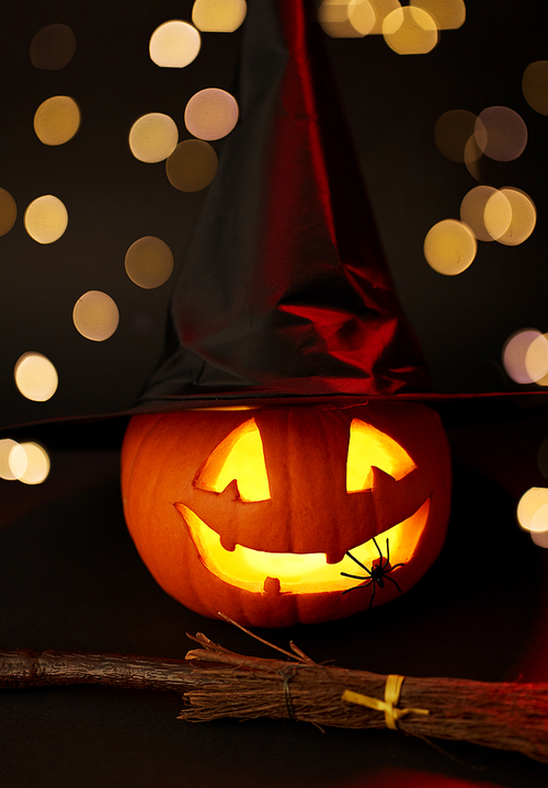 halloween and holiday decorations concept - jack-o-lantern in witch's hat with spider and broom in darkness over festive lights