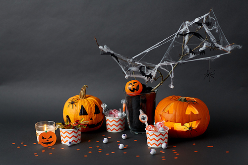 halloween and holiday decorations concept - jack-o-lantern or carved pumpkin, candies in paper cups, bats and spiders on spiderwab