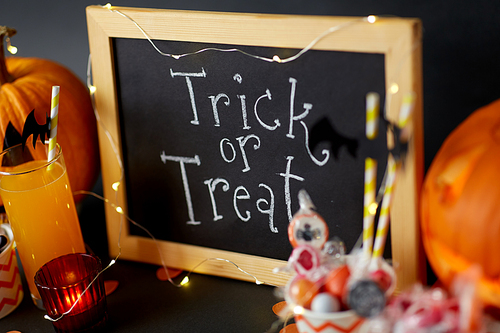 halloween and holiday decorations concept - close up of chalkboard with trick or treat lettering, jack-o-lanterns or carved pumpkins, candies, burning candles and glass of juice with paper straw