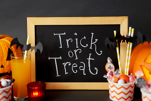 halloween and holiday decorations concept - chalkboard with trick or treat lettering, jack-o-lanterns or carved pumpkins, candies, burning candles and glass of juice with paper straw