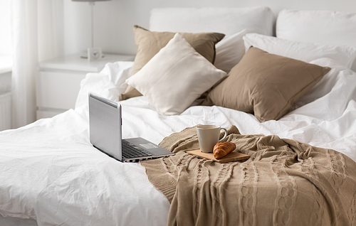 comfort, hygge and interior concept - laptop computer, cup of coffee and croissant on bed at cozy home
