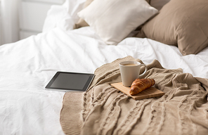 comfort, hygge and interior concept - tablet computer, cup of coffee and croissant on bed at cozy home