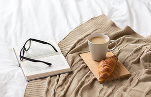 comfort, hygge and interior concept - coffee, croissant, blanket and book on bed at cozy home