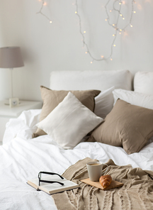 comfort, hygge and interior concept - coffee, croissant, blanket and book on bed at cozy home
