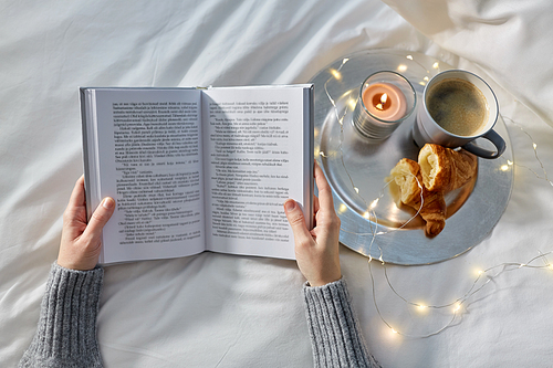 food, breakfast and people concept - hands of woman with book, coffee with croissant in bed